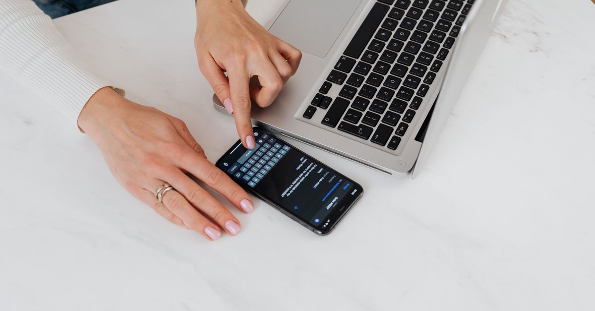 Close-up of hands checking a phone next to a laptop, the kind of session review that catches MFA bypass phishing